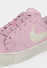 Κοντινό πλάνο ενός ροζ παπουτσιού Nike με κορδόνια στο ίδιο χρώμα, λευκό υφασμάτινο λογότυπο Swoosh και υποδήματα εκρού σε απλό φόντο.