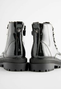 Bottes chevilles en cuir verni noir avec une texture brillante, des accents en forme d'étoile argentés, et des semelles en caoutchouc noir robuste. Conception à lacets.