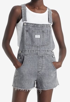 Personne portant une courte salopette en jean gris avec une poche avant et un ourlet effiloché, associée à un haut blanc sans manches, mains dans les poches.
