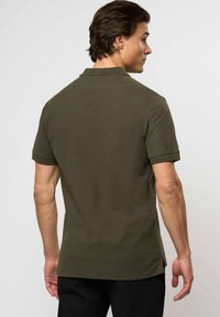 Muž s krátkými hnědými vlasy, oblečený v olivově zeleném polokošili s krátkým rukávem a černých kalhotách, stojí zády k pozadí světlejší barvy.