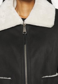 Närbild av en dragkedjefodrad jacka med en svart tygkropp och en fluffig vit krage, med en synlig halsringning som föreslår värme och stil.