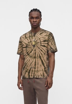 Mladý muž s vrkočmi, ktorý má na sebe hnedé a zelené tričko s tie-dye vzorom špirály a hnedé nohavice, stojaci pred bielym pozadím.