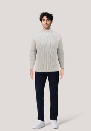 Maglione grigio chiaro a coste con collo alto, abbinato a pantaloni blu scuro e scarpe da ginnastica bianche, in piedi contro uno sfondo neutro.