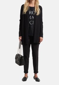 Cardigan in maglia nera sopra una t-shirt nera con grafica, pantaloni neri, mocassini neri e una borsa a tracolla nera trapuntata con catena.