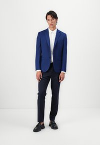 Blå blazer, hvit skjorte med knapper, mørke bukser og svarte sko. Blazeren har en enkelt knapp og en slank design med brystlomme.