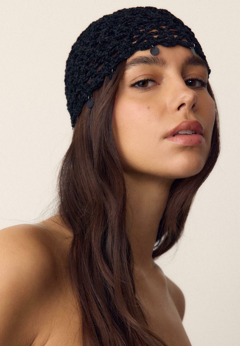 Jeune femme aux longs cheveux bruns foncés portant un foulard noir en crochet avec de petites décorations rondes, expression neutre, fond beige.