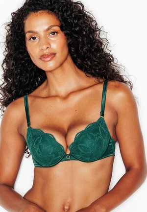 Donna con capelli scuri e ricci che indossa un reggiseno verde in pizzo e raso su sfondo bianco.