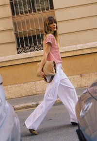 Femme en chemise rouge rayée et pantalon blanc large marchant dans une rue de la ville, tenant une grande pochette beige, avec des voitures garées à proximité.