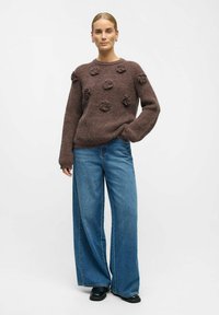 Rudas megztinis su tekstūriniais žiedais, derinamas su laisvais mėlynais džinsais ir juodais batais; kasdieniškas kirpimas ir atsipalaidavęs dizainas.