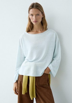 Kobieta o długich blond włosach ma na sobie luźny, jasnoniebieski sweter i brązowe spodnie, z splecionym wokół talii żółtym swetrem, stoi przy białej ścianie.