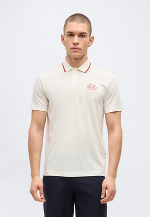 Rövid hajú férfi fehér, piros szegéllyel és EA7 logóval ellátott pólóingben, valamint sötét nadrágban, egyszerű fehér háttér előtt áll.