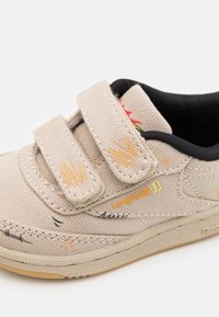 Beige canvas sneaker med kardborreband, svart foder och färgglada broderade detaljer. Texturerad yttersula med orange accenter.