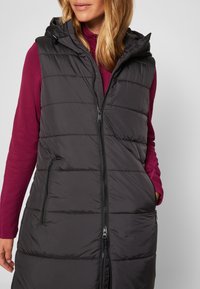 Femme portant un gilet matelassé noir sans manches avec capuche par-dessus un haut violet à manches longues, debout avec les mains partiellement dans les poches du gilet.