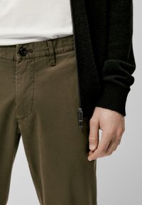 Donker gebreide cardigan met een rits, gecombineerd met khaki broeken met een knoop en zijzakken. Close-up van een hand op de stof.