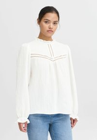 Witte blouse met lange mouwen en ruches bij de kraag, van gestructureerde stof, met verticale stiksels op de borst. Gecombineerd met blauwe jeans.