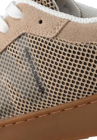 Tan mockaskinn och mesh-sneakers med vita snören, en strukturerad brun sula och framträdande branding på sidan i svart.