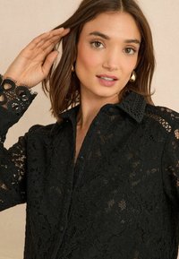 Camicia nera in pizzo con colletto testurizzato e intricati motivi floreali; presenta chiusura a bottoni e polsini accentuati in pizzo.