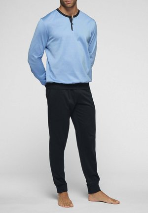 Uomo in piedi a piedi nudi che indossa una maglia henley a maniche lunghe celeste con rifiniture nere e pantaloni jogger neri aderenti su sfondo semplice.