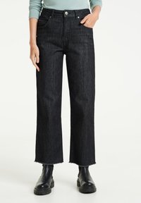 Osoba oblečená v černých širokých džínách s vysokým pasem a roztrhanými lemy, černých kotníčkových botách, ruka v kapse, viditelný světle modrý dlouhý rukáv.