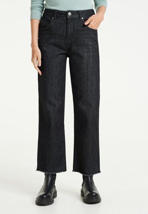 Personne portant un jean large taille haute noir avec des ourlets bruts et des bottines noires, main dans la poche, haut à manches longues bleu clair visible.