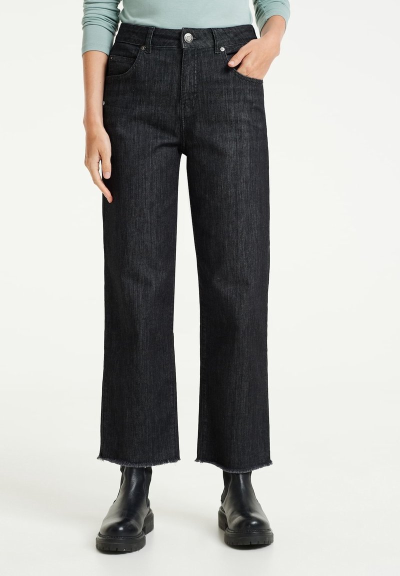 Osoba oblečená v černých širokých džínách s vysokým pasem a roztrhanými lemy, černých kotníčkových botách, ruka v kapse, viditelný světle modrý dlouhý rukáv.