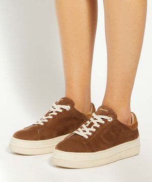 Jambes portant des baskets en daim marron avec des lacets blancs et des semelles blanches épaisses, debout sur une surface blanche.