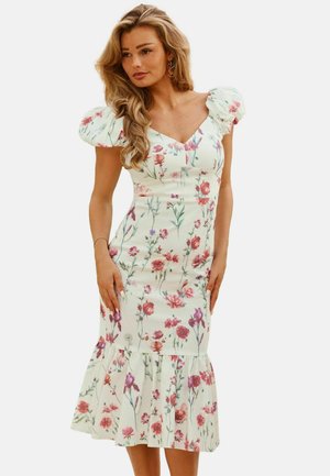 Kobieta w białej sukience midi z różowo-fioletowym kwiatowym wzorem, z bufkami, dopasowaną talią i falbaną na dole, stojąca na jednolitym tle.