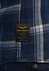 Blauwe geruite overhemd met marineblauwe, lichtblauwe en witte patronen. Heeft een zwart label met gouden letters en hardware-accenten op de zak.