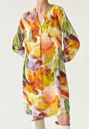 Luźna sukienka z długim rękawem w akwarelowy kwiatowy wzór w odcieniach pomarańczowego, zielonego, żółtego i fioletowego, noszona z białymi kozakami za kolano.