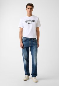 T-shirt bianca in cotone con logo nero "GUESS" e dettaglio "EST. 1981", abbinata a jeans denim blu con accenti sbiaditi e sneakers bianche.