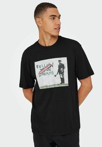 Camiseta de algodón negra con un estampado gráfico de una persona frente a una pared con el texto "SIGUE TUS SUEÑOS" y un sello rojo de "CANCELADO".