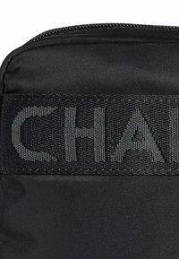 Bolsa de tela negra con un logotipo bordado texturizado en gris oscuro. Superficie suave con un cierre de cremallera en la parte superior.