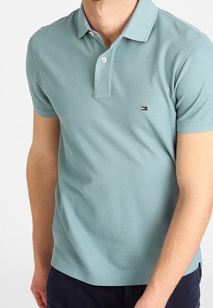 Homme portant un polo bleu clair à manches courtes avec un petit logo rouge, blanc et bleu sur la poitrine, associé à un pantalon foncé.