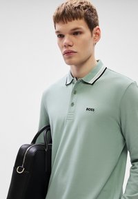 Polo verde claro com gola preta e branca, apresentando um pequeno logótipo preto "BOSS". O modelo segura uma mala de couro preta.