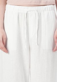 White linen pants with an elastic waistband, featuring a drawstring detail and side pockets. Smooth texture and loose fit.