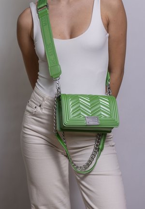 Personne portant un débardeur blanc et un pantalon beige clair avec un sac bandoulière matelassé vert vif présentant des détails de chaîne et de sangle argentés.