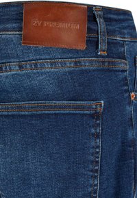 Jeans en denim bleu foncé avec une étiquette en cuir marron portant l'inscription "2Y PREMIUM" au niveau de la taille, présentant des coutures orange et un détail de poche.