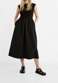 Abito senza maniche nero con un corpetto attillato e una gonna fluida, realizzato in tessuto leggero. Abbinato a sandali neri con fasce.