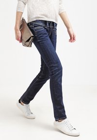 Jeans ajustados de color azul oscuro con una cintura de altura media, costuras en contraste y un acabado suave. Combinados con un suéter claro y zapatillas blancas.