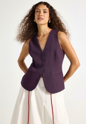 Gilet viola senza maniche con profondo scollo a V, chiusura frontale con bottoni e due tasche laterali. Indossato sopra una gonna bianca con accenti rossi.
