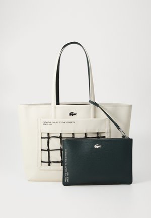 Torba za nakupovanje v off-white barvi, ki ima vzorec črtaste mreže in vezen logotip. Vključuje snemljivo temno zeleno torbico z teksturirano površino in srebrnimi detajli.
