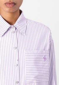 Lavender striped shirt with a pocket, featuring silver heart-shaped buttons and a small logo embroidered in purple on the pocket.