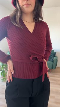 Sweater envolvente em bordeaux com textura canelada e mangas longas, com um laço na cintura, combinado com calças pretas. O sweater apresenta riscas verticais e um decote em V.