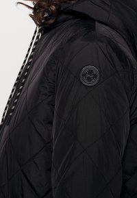 Chaqueta de plumas negra acolchada con textura brillante, que presenta cordones de contraste y un parche de logo redondo en la manga izquierda.