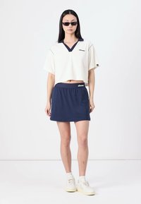 T-shirt court blanc avec des accents navy, associé à une jupe à taille élastique navy. Détails logo en tissu. Le mannequin porte des lunettes de soleil et des sneakers.
