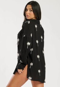 Camisa negra de tejido ligero, con bordados de palmeras plateadas en toda la prenda, corte ajustado con cuello abotonado y aberturas laterales.