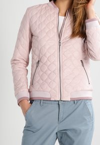 Frau trägt eine hellrosa gesteppte Bomberjacke mit Reißverschlusstaschen und hellblauen Hosen, Hand in der Tasche, vor weißem Hintergrund.