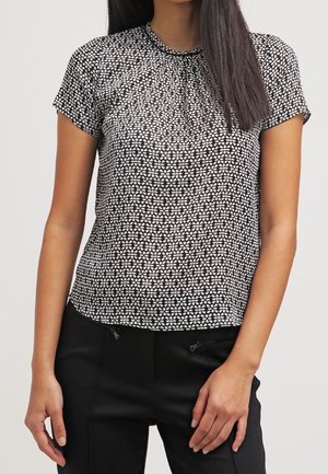Vrouw die een zwart-wit blouse met korte mouwen en een patroon draagt, en zwarte broek, staand tegen een egale achtergrond.