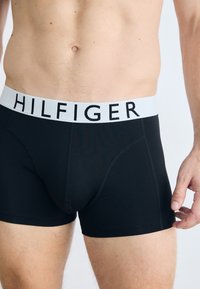 Schwarze Boxershorts mit einem weißen Gummibund, der das Logo "HILFIGER" trägt. Glattes Material, enganliegendes Design und Mid-Thigh-Länge.