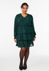 Abito a balze verde scuro con maniche lunghe, scollatura a V, motivo floreale testurizzato e vita arricciata. Indossato con ballerine nere decorate.